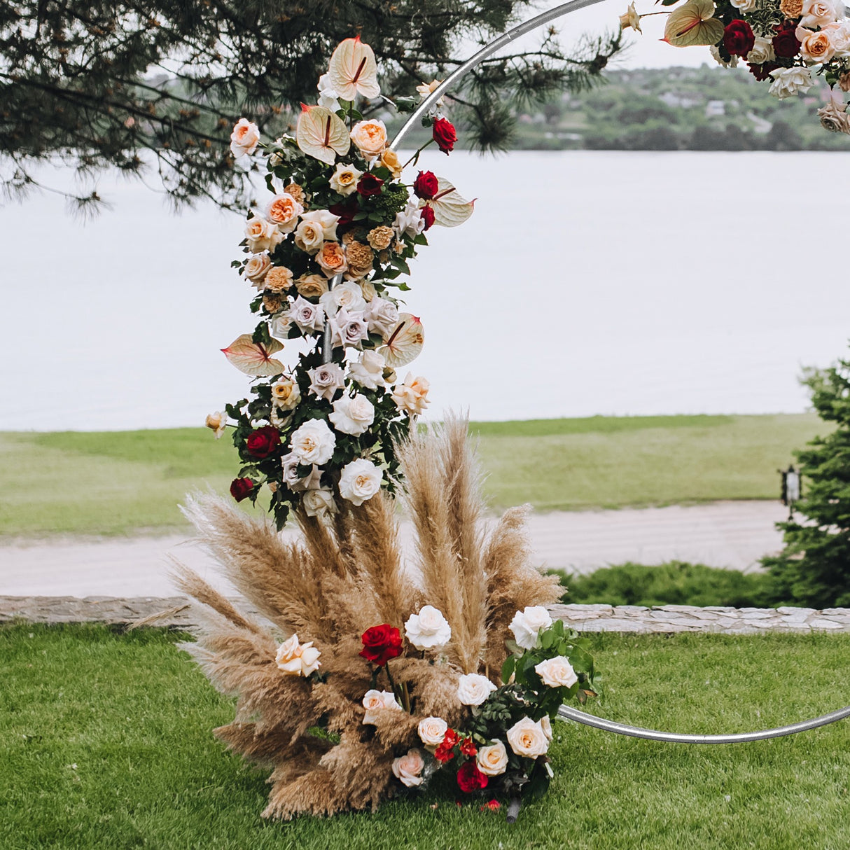 Arco decorativo per matrimoni in argento, adornato con rose bianche, rosse e fiori di pampas, su sfondo verde e acqua