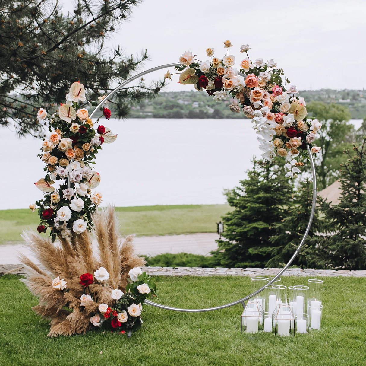 Arco decorativo per matrimoni in argento, adornato con fiori colorati e pampas, circondato da candele bianche su prato verde