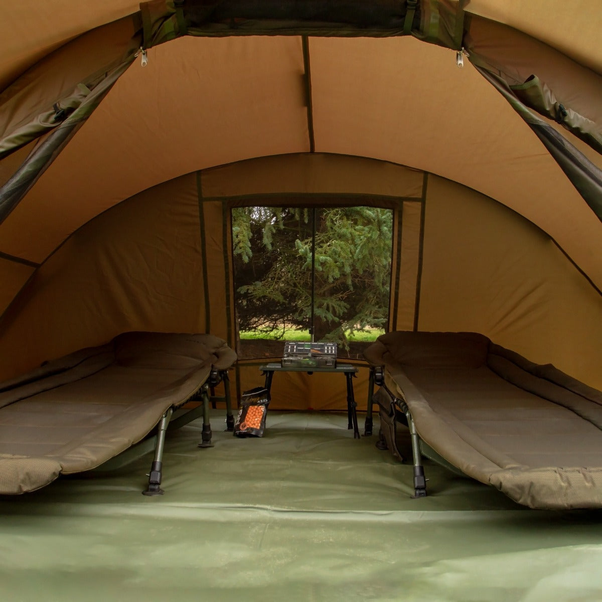 Interno di una tenda bivvy da pesca per due persone, con letti pieghevoli verdi e un tavolo al centro, pareti marroni e vista su alberi verdi