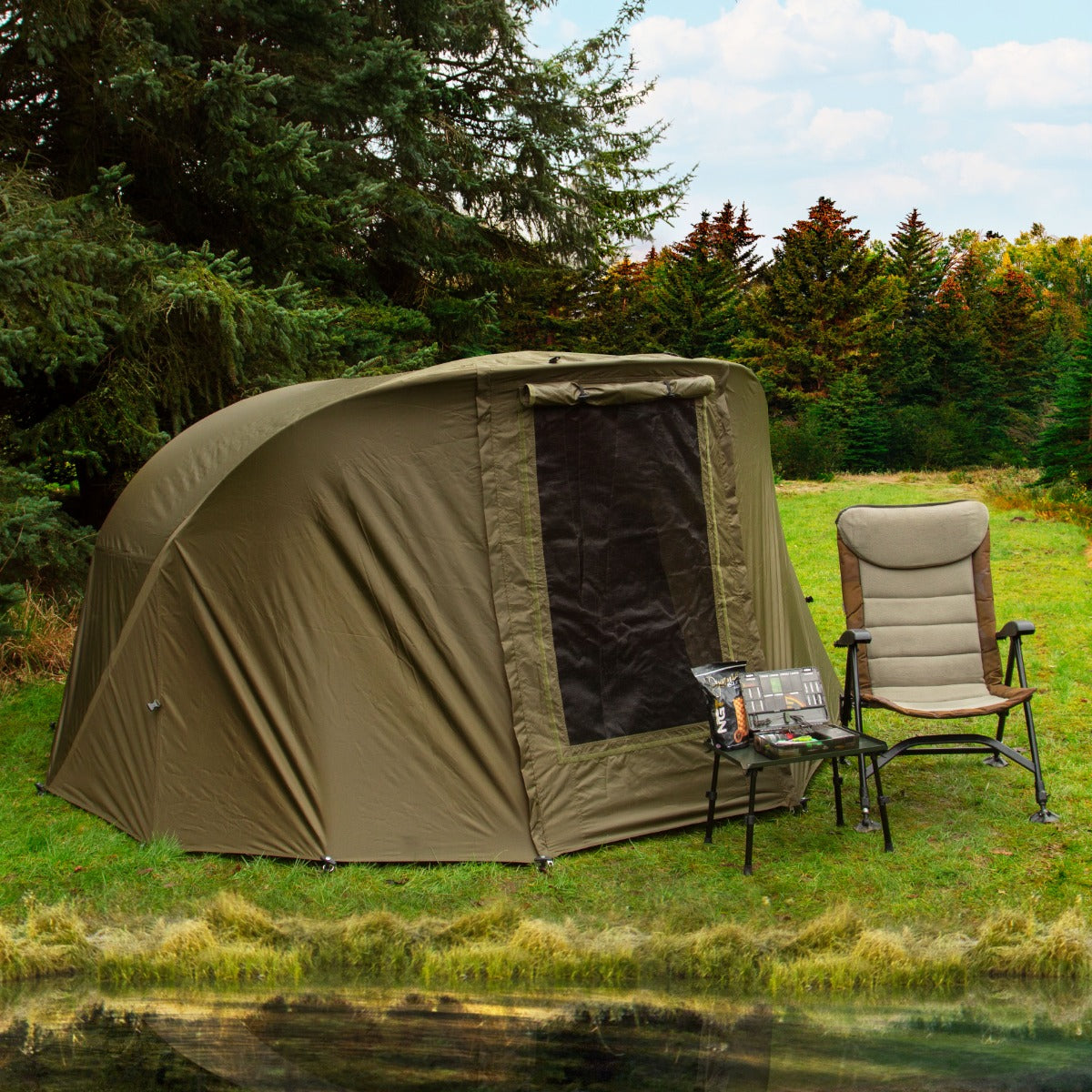 Tenda bivvy da pesca verde oliva per due persone, con sedia pieghevole accanto e vista su un prato verde e alberi sullo sfondo