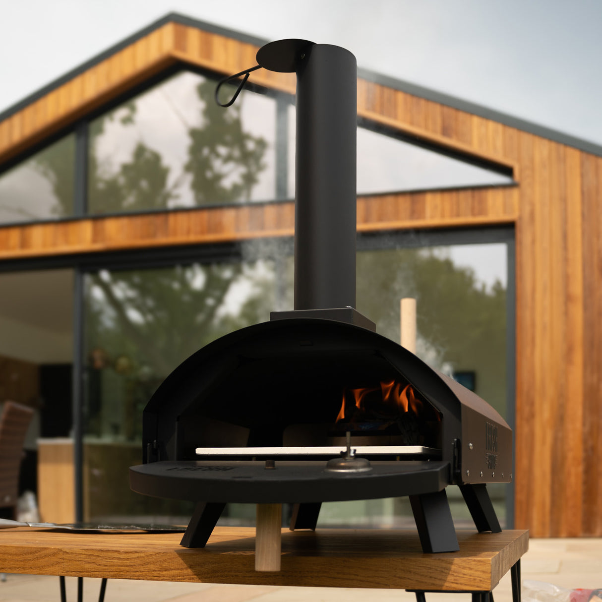 Forno per pizza da esterno Fresh Grills Classic, struttura nera, camino alto, fiamma visibile, posizionato su un tavolo di legno, sfondo moderno