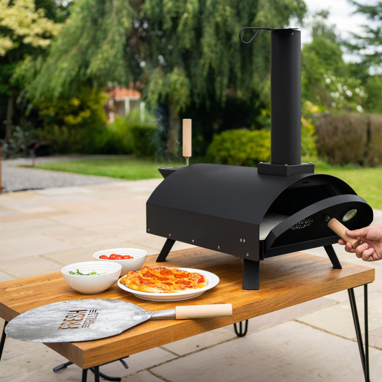 Forno per pizza da esterno Fresh Grills Classic, struttura nera, camino verticale, accanto a pizza e ingredienti freschi su un tavolo di legno