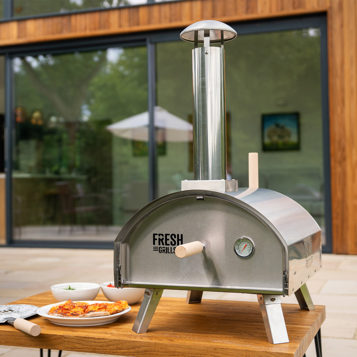 Forno per pizza da esterno Fresh Grills Premium in acciaio inox, con camino, su tavolo di legno, circondato da piatti di pizza e condimenti