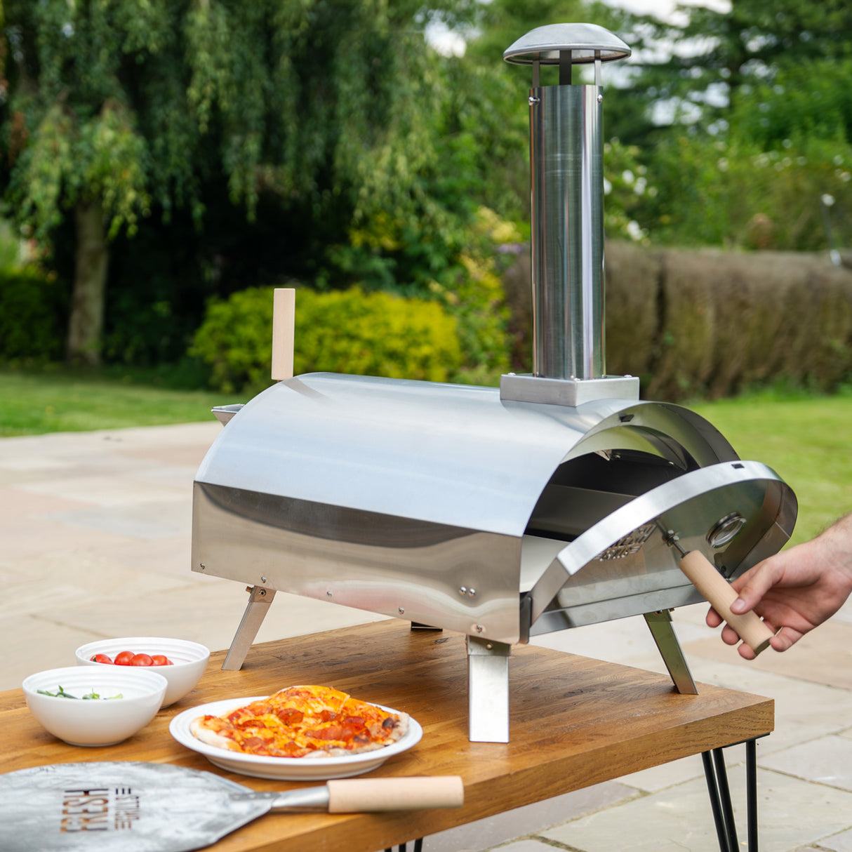 Forno per pizza da esterno in acciaio inox lucido, con camino, posizionato su un tavolo di legno accanto a pizze e ingredienti freschi