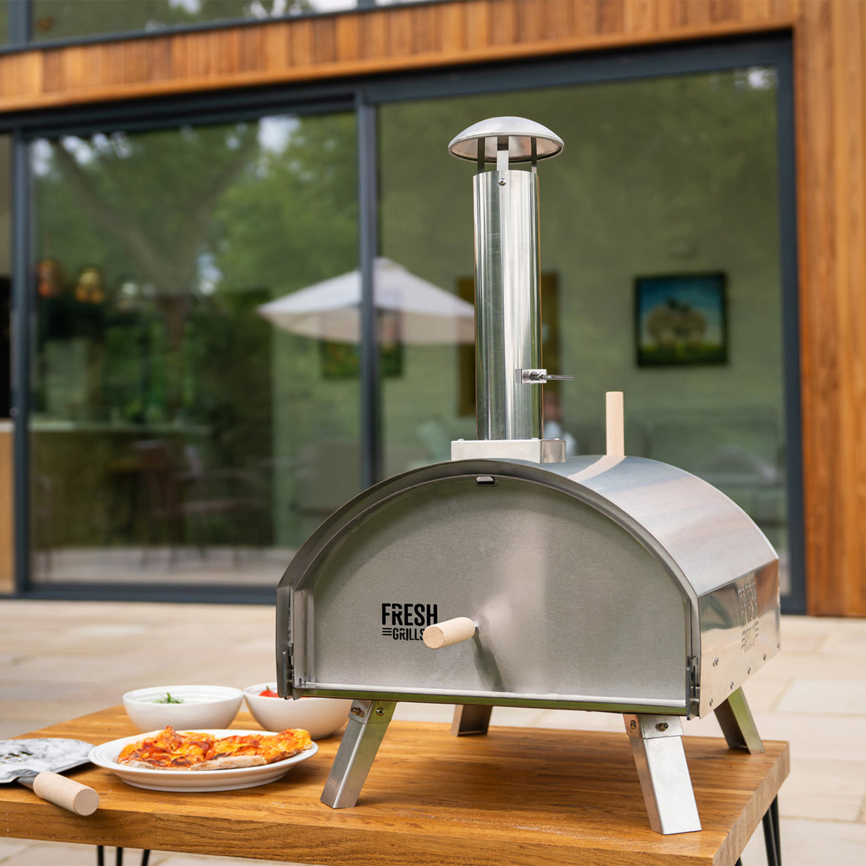 Forno per pizza all'aperto Fresh Grills Extra Large in acciaio inox, con camino, su tavolo di legno, circondato da piatti di condimenti freschi
