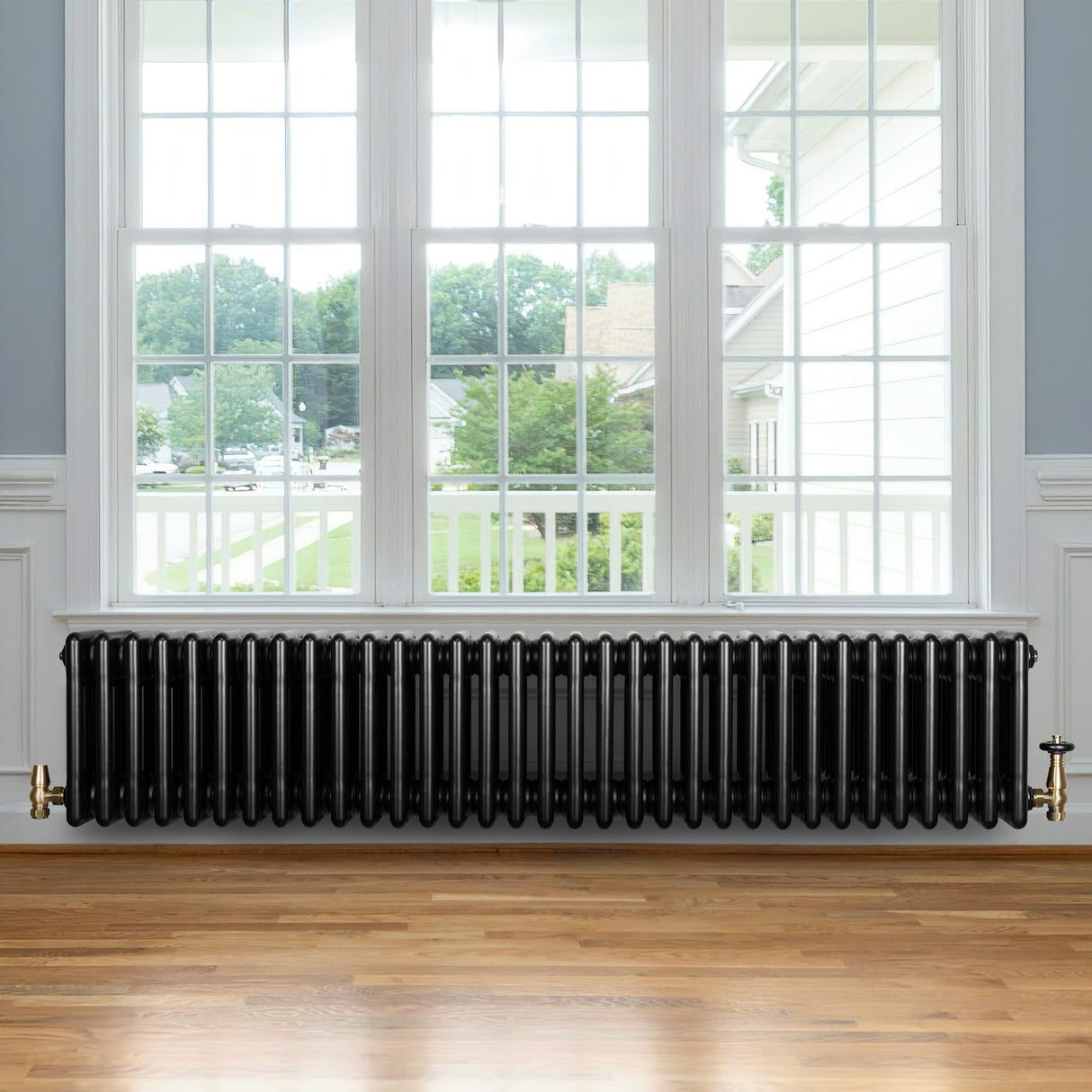 Radiatore tradizionale a 4 colonne, finitura nera lucida, posizionato sotto una finestra con vista su un giardino, su pavimento in legno chiaro