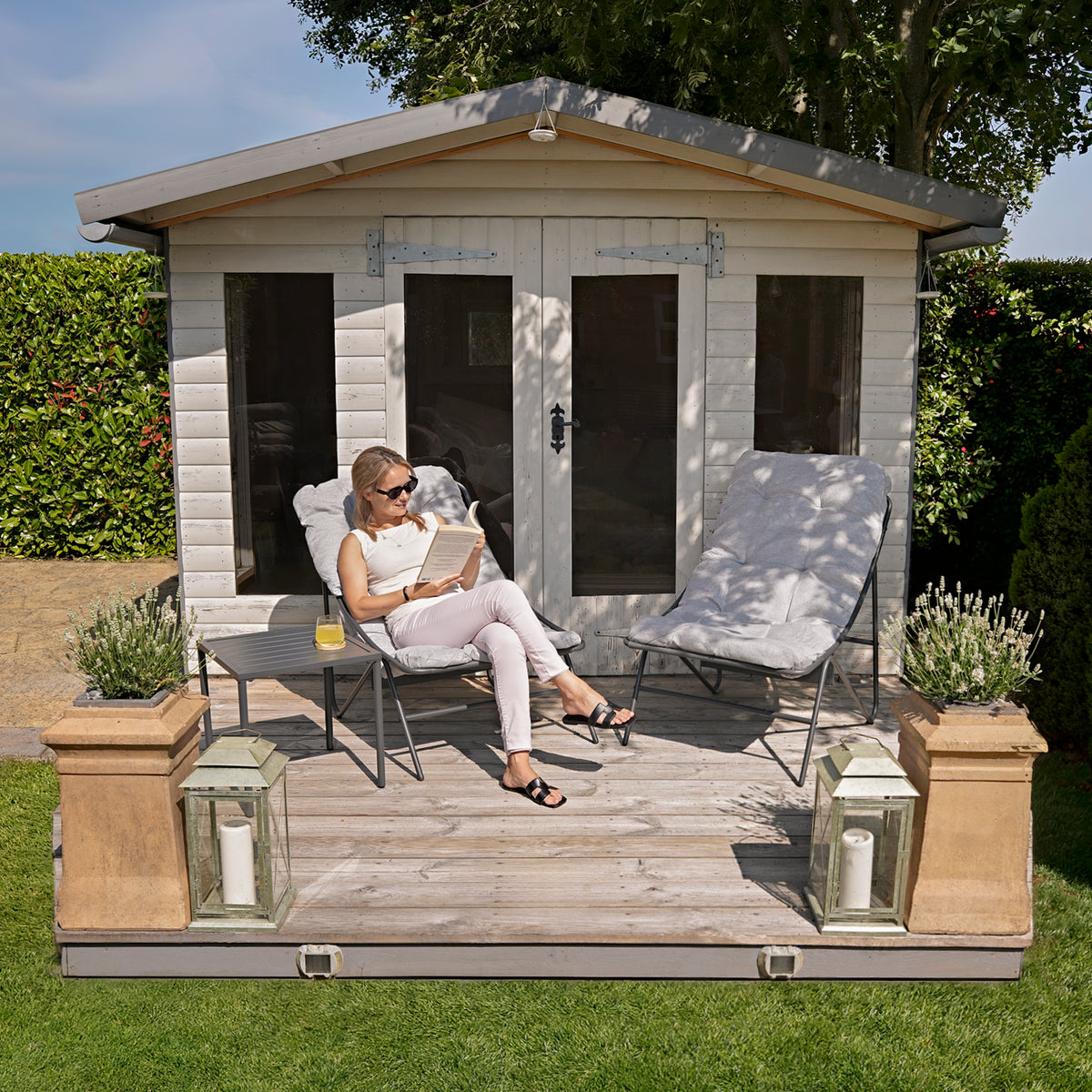 Una donna seduta su un patio in legno, legge un libro, circondata da piante verdi e un elegante casetta bianca in legno