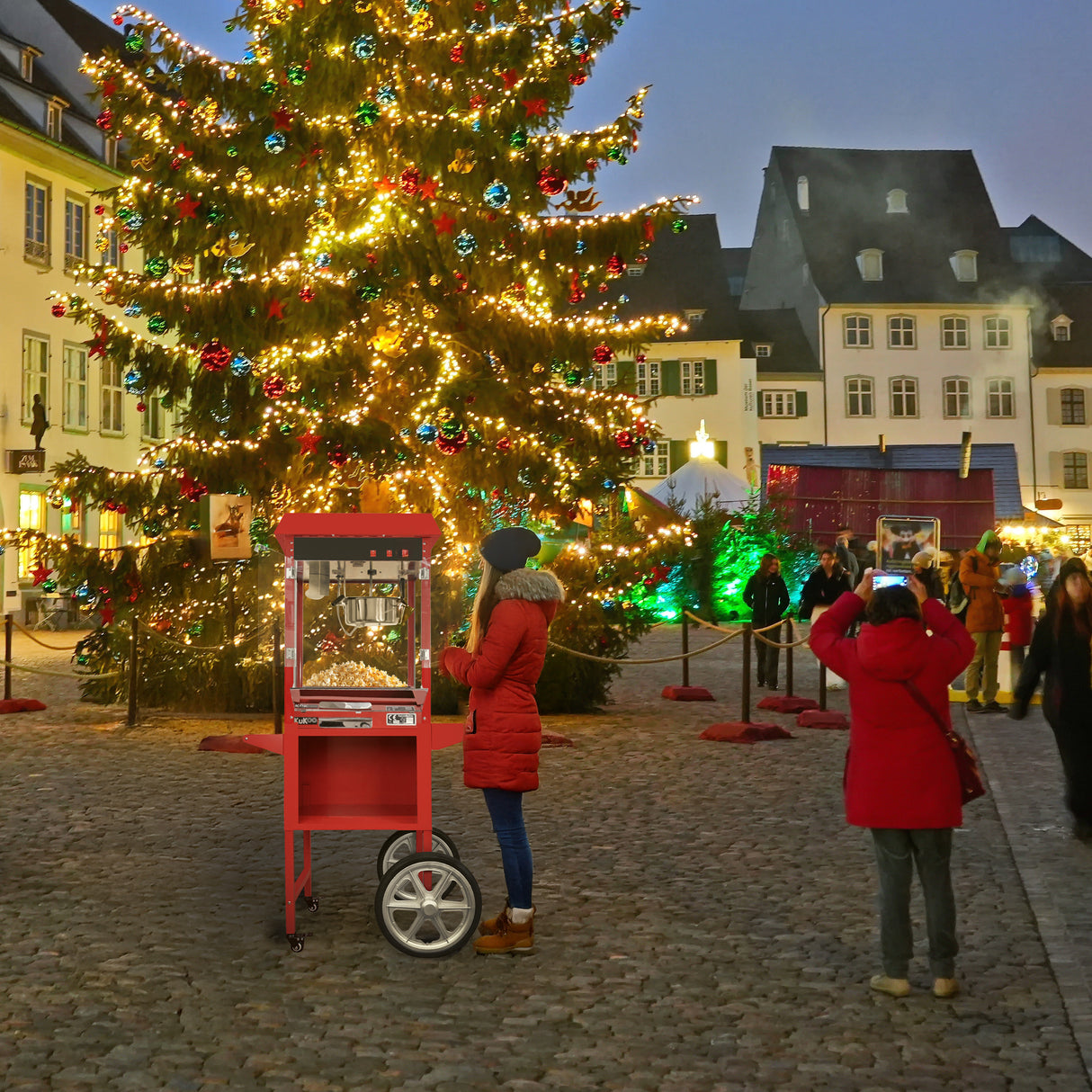 Carrello per popcorn KuKoo rosso brillante, con ruote, posizionato davanti a un albero di Natale decorato in una piazza affollata
