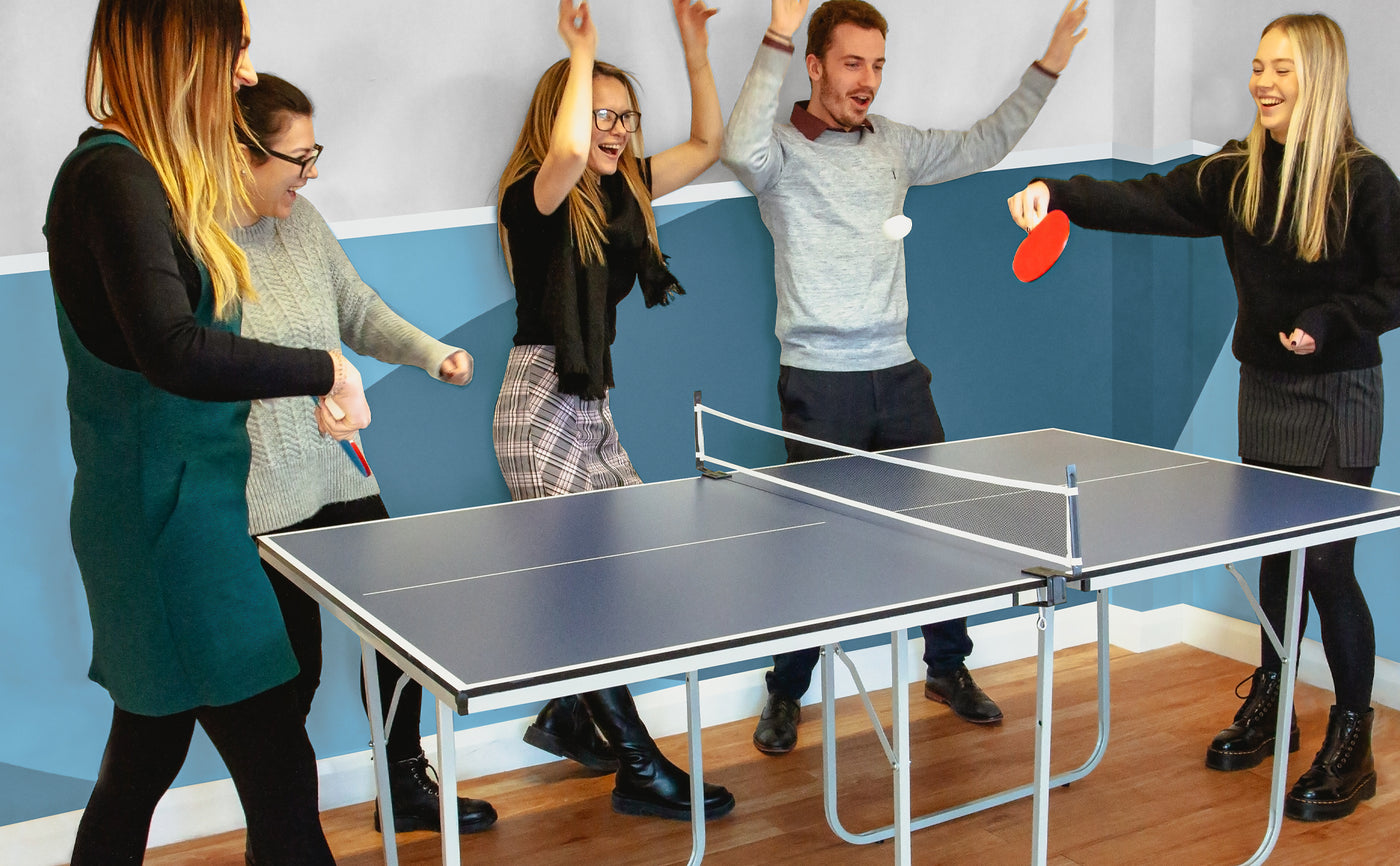 Gruppo di cinque persone che si divertono giocando a ping pong, con tavolo blu e pareti colorate, espressioni di gioia e entusiasmo