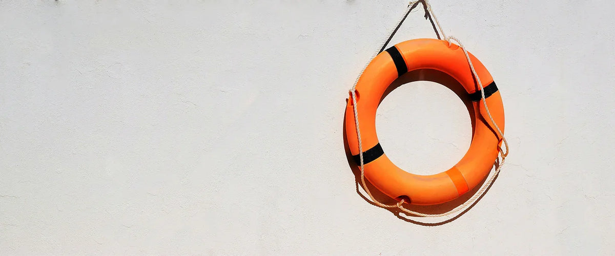 Salvagente arancione brillante appeso a una parete bianca, con strisce nere e corda di fissaggio, simbolo di sicurezza in acqua