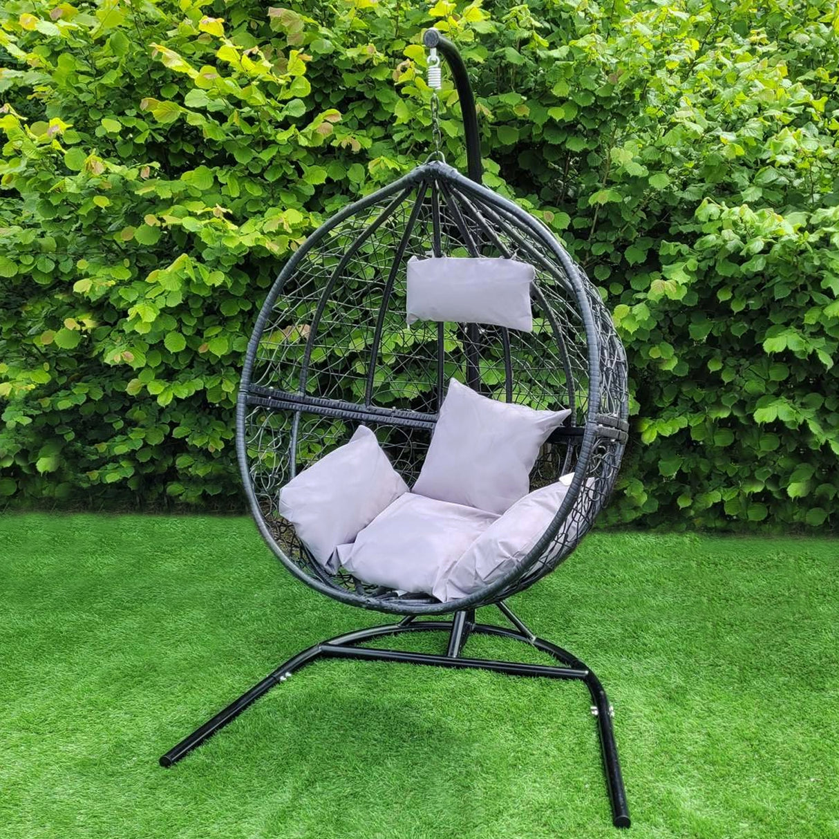 Sedia sospesa a uovo in rattan nero con cuscini grigi, appesa a un supporto metallico su prato verde, ideale per esterni