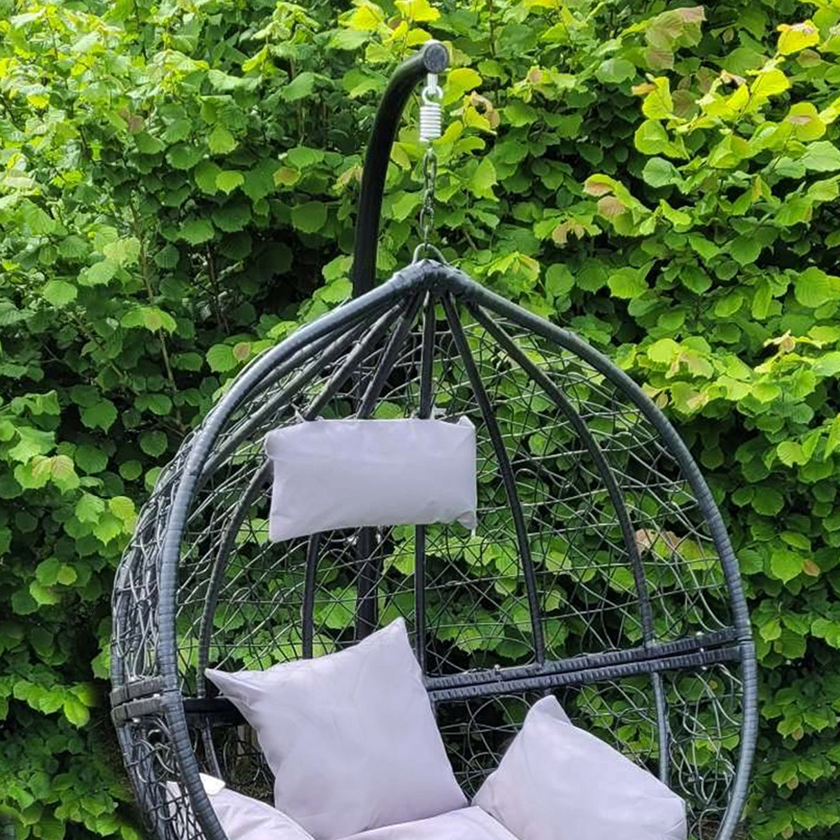 Sedia sospesa a uovo da giardino in rattan nero, con cuscini bianchi, appesa a un supporto elegante tra foglie verdi