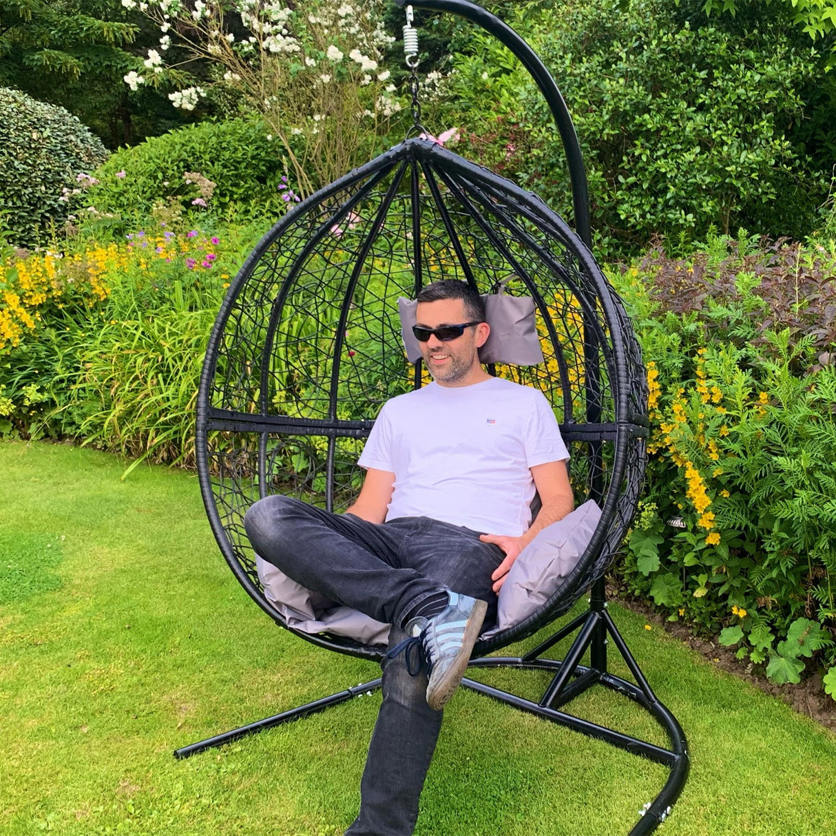 Sedia dondolo sospesa a forma di uovo in rattan grigio, con cuscini morbidi, posizionata in un giardino fiorito verde brillante
