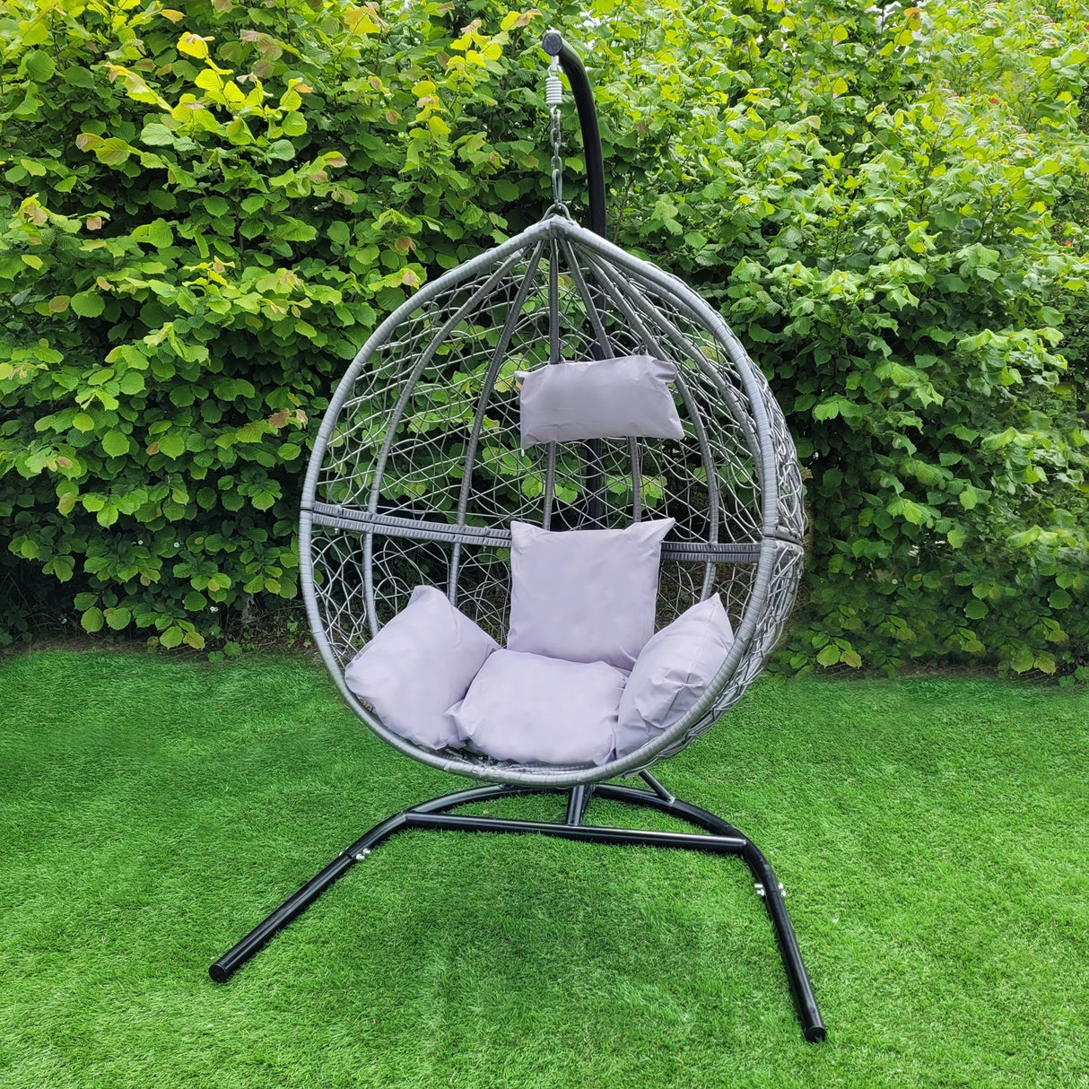 Sedia dondolo sospesa a uovo in rattan grigio con cuscini morbidi, appesa a un supporto nero, immersa nel verde del giardino
