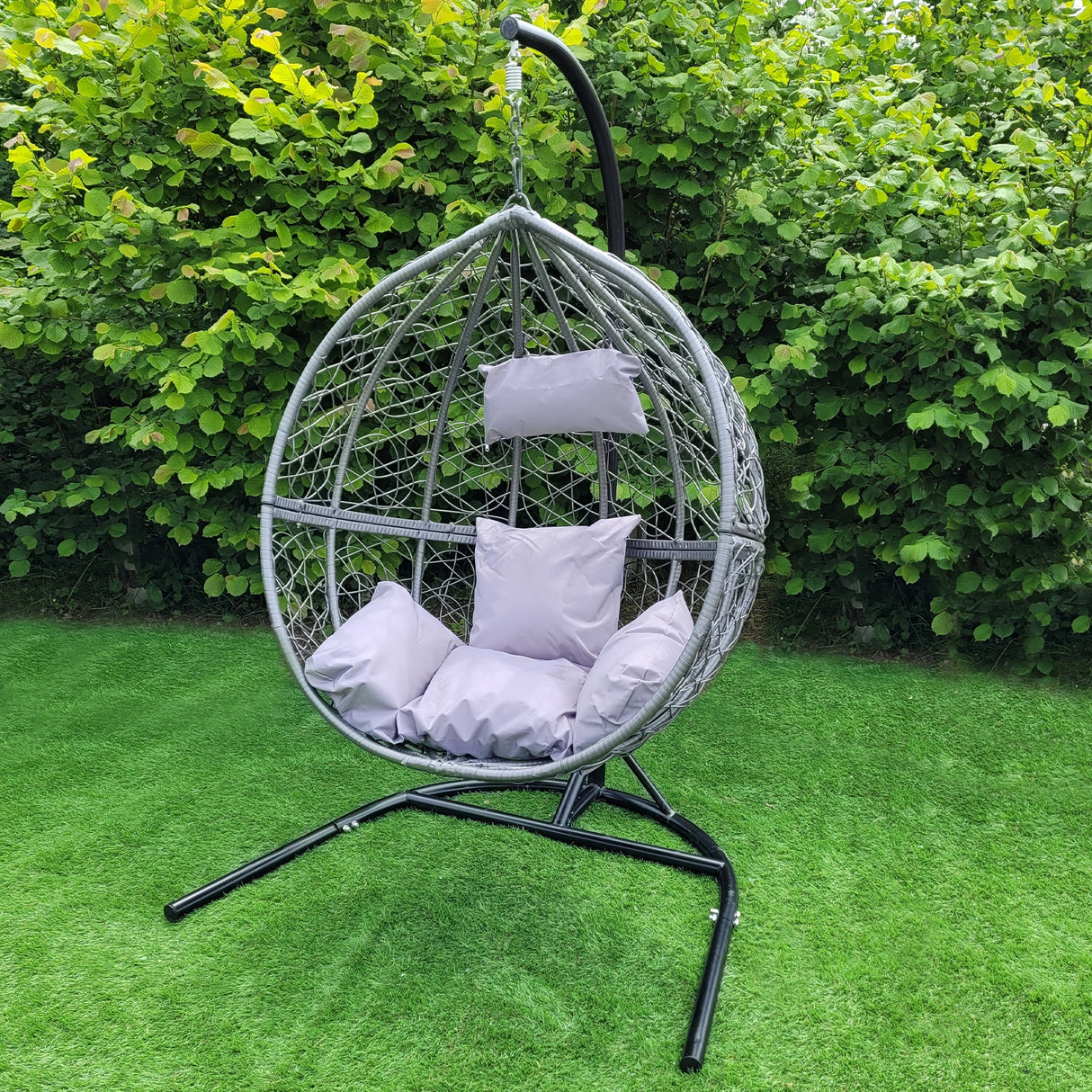 Sedia sospesa a uovo da giardino in vimini rattan grigio, con cuscini morbidi e struttura nera, immersa nel verde del giardino