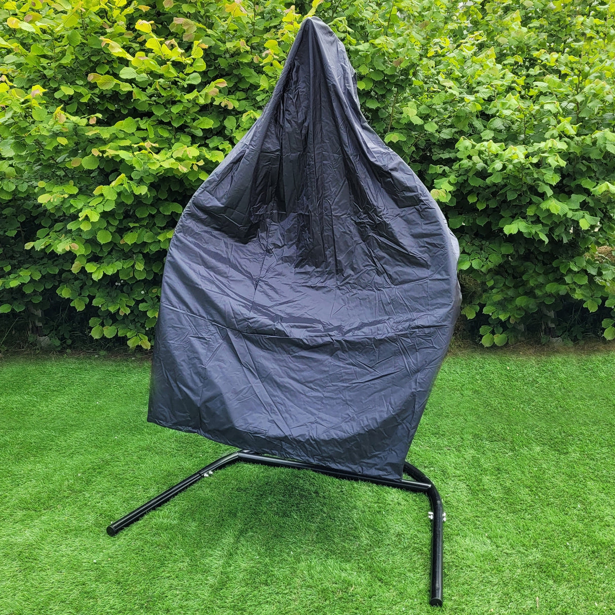 Sedia sospesa a uovo da giardino in rattan grigio, coperta da un telo protettivo, posizionata su un supporto nero in un giardino verde