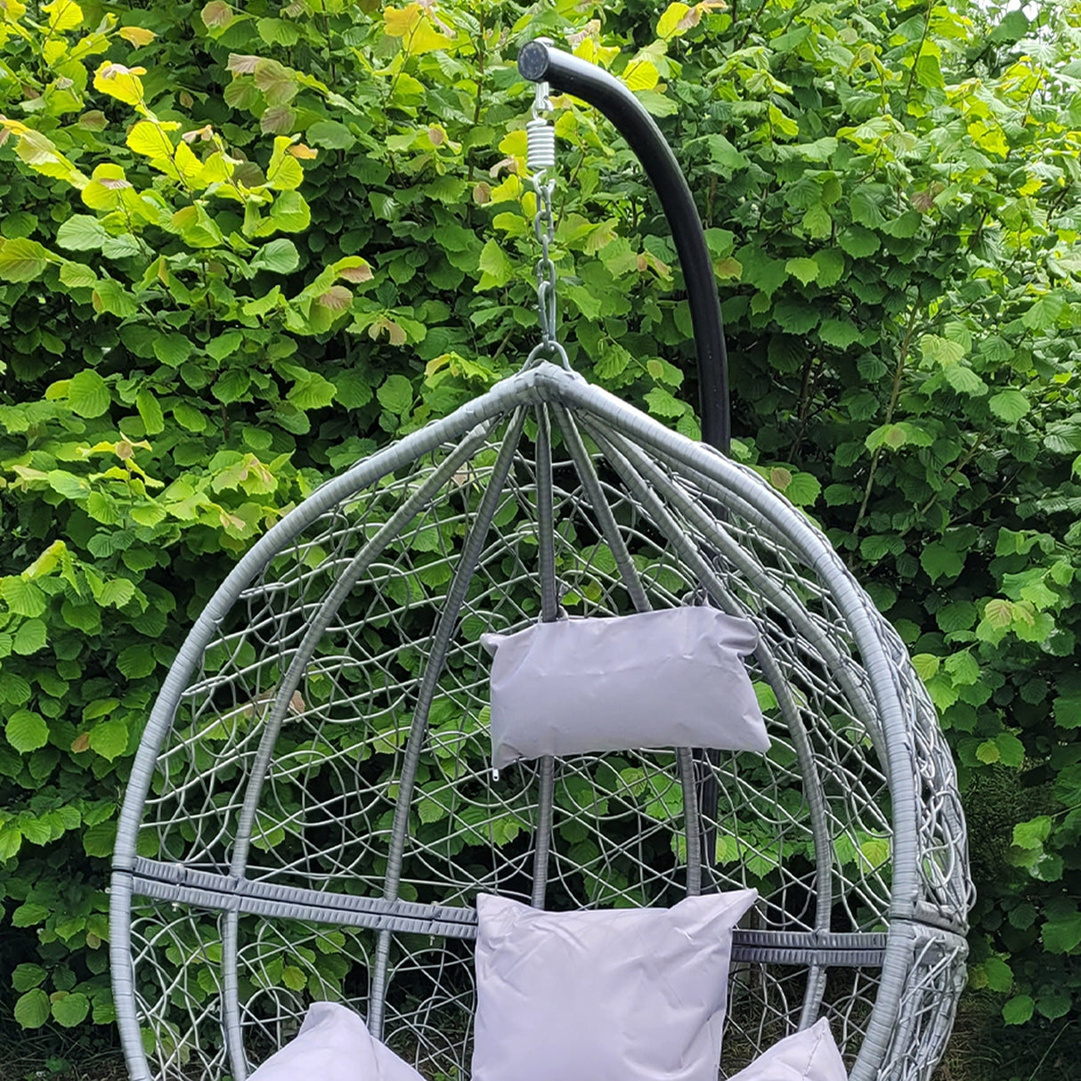 Sedia sospesa a uovo in rattan grigio con cuscini morbidi, appesa a un supporto nero, immersa in un verde giardino lussureggiante