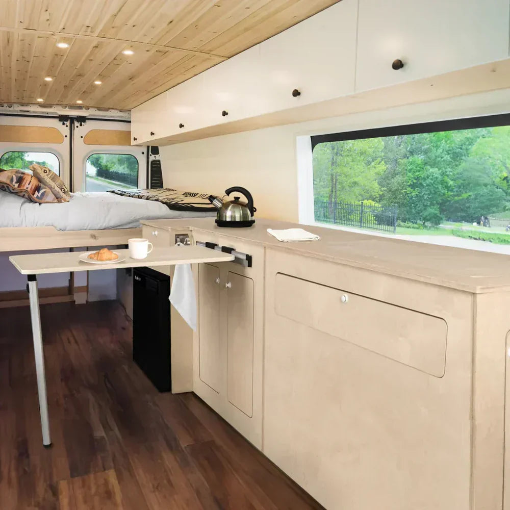 Interno di un camper van con cucina in legno chiaro, letto matrimoniale, tavolo pieghevole e vista su un paesaggio verde all'esterno