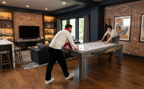 Due tavoli da gioco in legno grigio, uno con piani di gioco per hockey e biliardo, in un soggiorno moderno con mattoni a vista