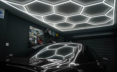 Illuminazione LED esagonale bianca su soffitto scuro di un garage, riflessi sul cofano di un'auto nera, atmosfera moderna e accogliente