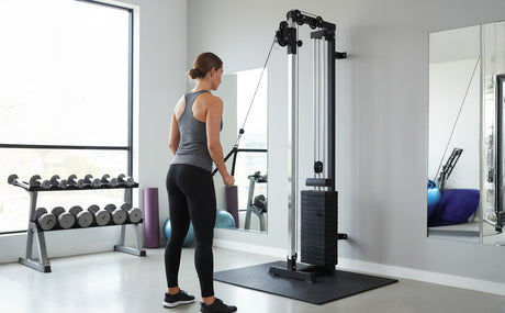 Donna che utilizza una macchina a cavi da parete in palestra, indossando top grigio e leggings neri, con attrezzi sullo sfondo