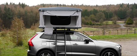Tenda da campeggio montata su un'auto grigia, con struttura in metallo, tessuto grigio e sfondo di boschi verdi e colline