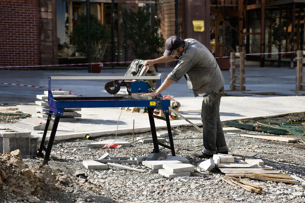 Lavoratore utilizza un tagliapiastrelle ad acqua blu su un cantiere, circondato da materiali da costruzione e pavimentazione in pietra