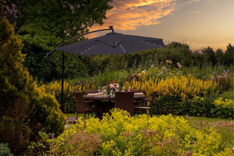 Ombrellone da giardino nero con luci, posizionato su un tavolo da pranzo circondato da fiori gialli e verde lussureggiante al tramonto