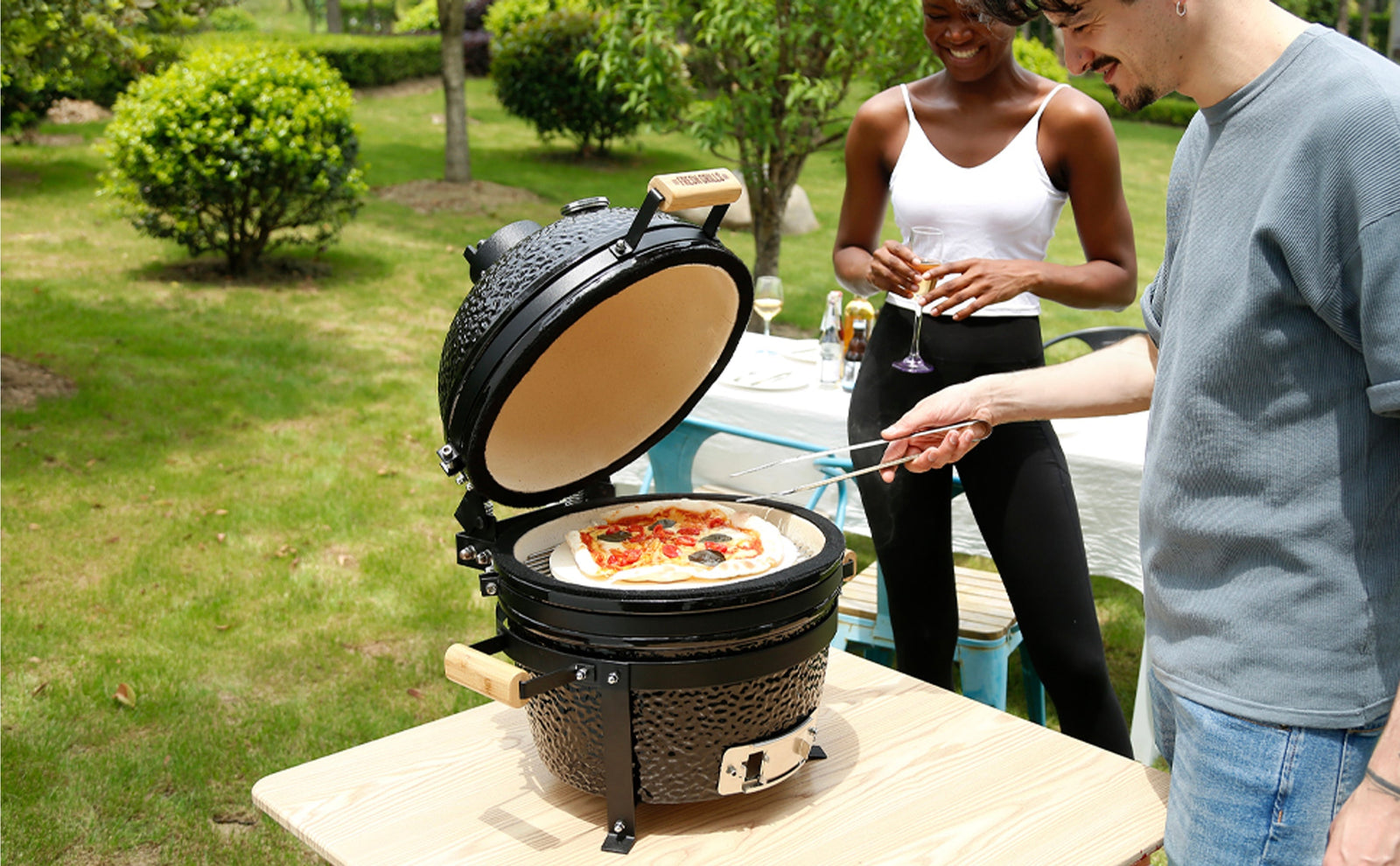 Grill Kamado nero in giardino, con pizza colorata e ingredienti freschi, mentre due persone sorridono e brindano accanto