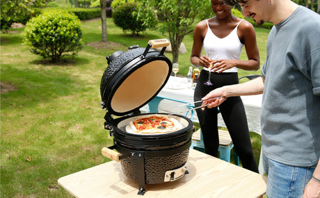 Grill Kamado nero in giardino, con pizza colorata e ingredienti freschi, mentre due persone sorridono e brindano accanto