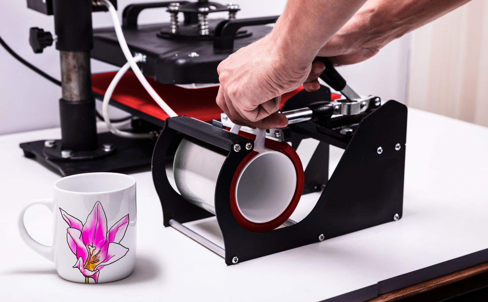 Pressa per tazze a sublimazione con manico rosso, accanto a una tazza bianca decorata con un'orchidea rosa su sfondo chiaro
