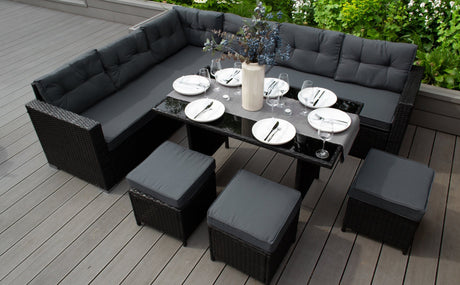 Set da pranzo in rattan con cuscini grigi, tavolo nero lucido e decorazione floreale, circondato da un giardino verde lussureggiante
