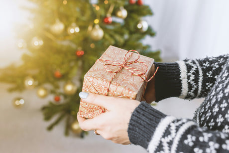 Scatola regalo avvolta in carta dorata con motivo, tenuta da mani in maglione nero, davanti a un albero di Natale decorato