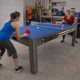 Tavolo multigioco 7-in-1 grigio con superficie blu, due persone che giocano a ping pong, ambiente di garage ben organizzato