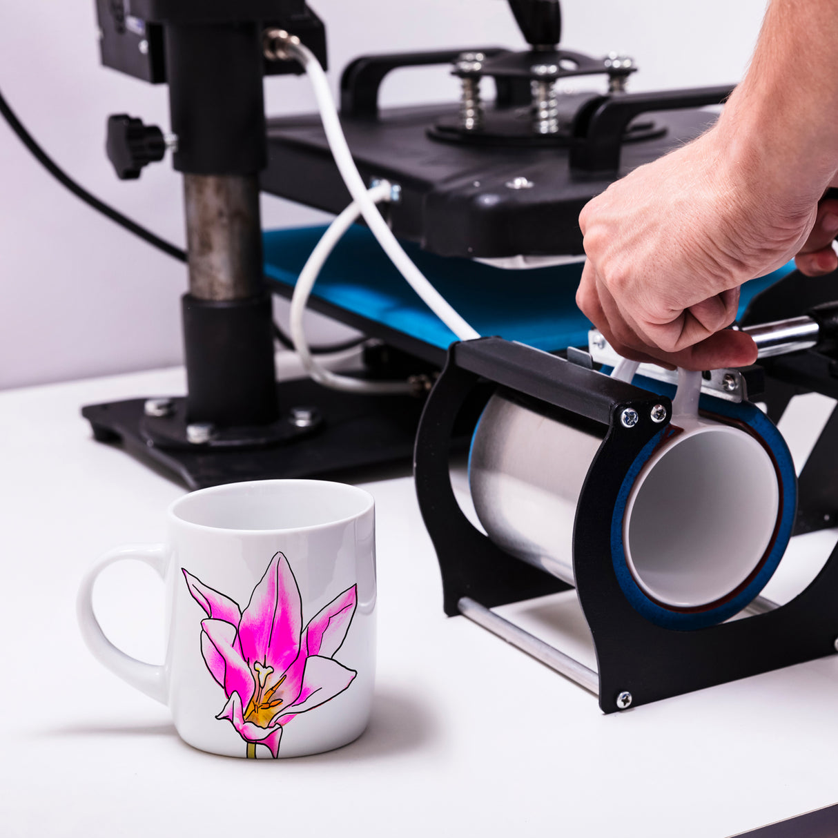 Tazza in ceramica bianca con un disegno di orchidea rosa, accanto a una pressa per sublimazione su sfondo neutro