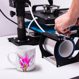 Tazza in ceramica bianca con un disegno di orchidea rosa, accanto a una pressa per sublimazione su sfondo neutro