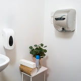 Asciugamani elettrico Maxblast grigio montato a parete, dispenser di sapone bianco e pianta verde su tavolino in un bagno moderno