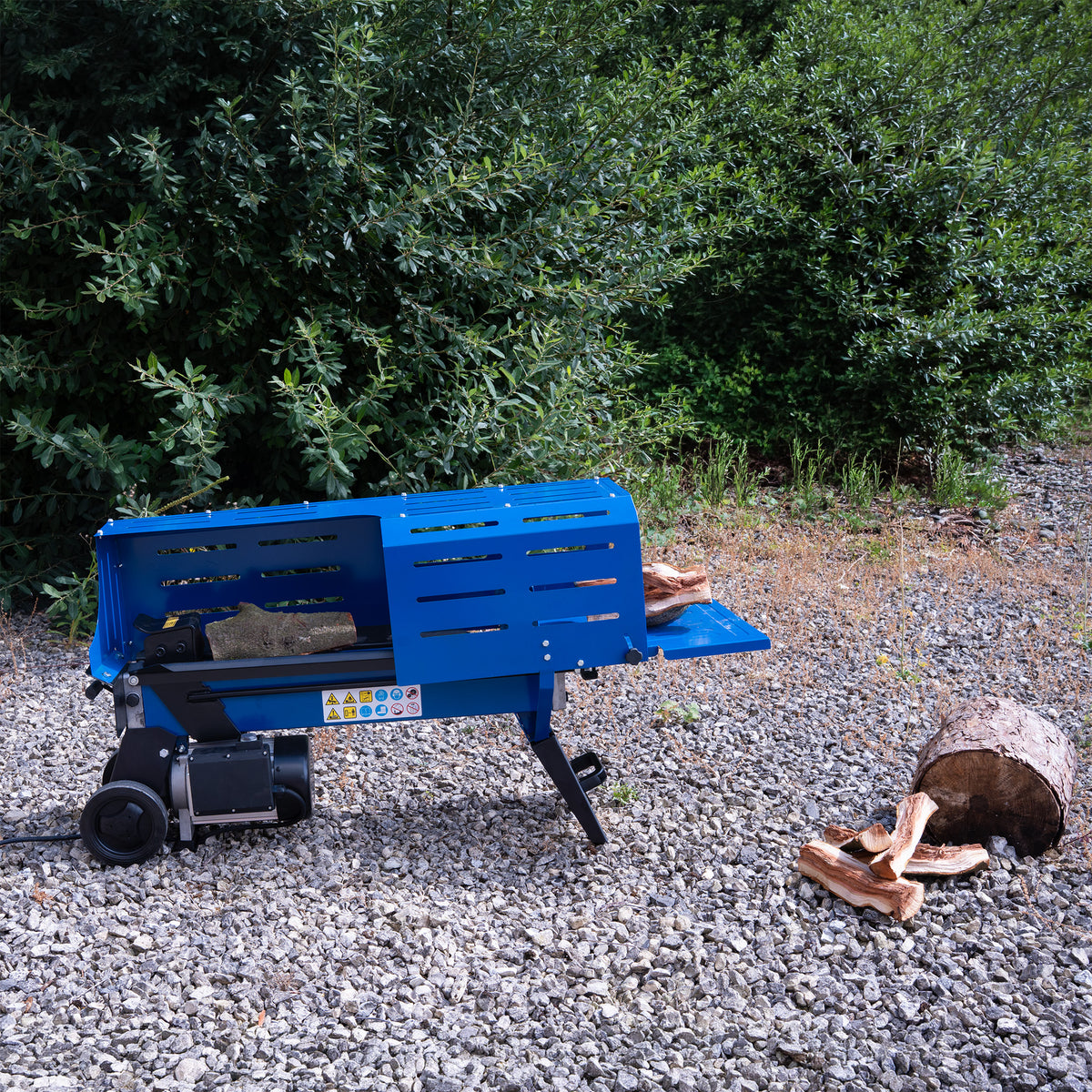 Spaccalegna elettrico blu su terreno ghiaioso, con tronchi di legno accanto e vegetazione verde sullo sfondo