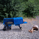 Spaccalegna elettrico blu su terreno ghiaioso, con tronchi di legno accanto e vegetazione verde sullo sfondo