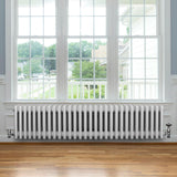 Radiatore tradizionale a 4 colonne, bianco, lungo 1370 mm, posizionato sotto una finestra con vista su un giardino, su pavimento in legno chiaro