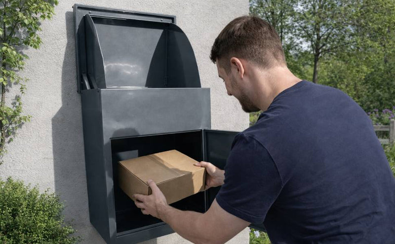 Uomo che inserisce un pacco marrone in una cassetta postale grigia, con un design moderno e una finitura liscia, circondato da verde