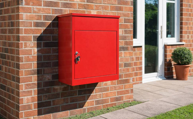 Cassetta postale rossa brillante montata su un muro di mattoni, con una chiave appesa e un vaso di piante accanto alla porta
