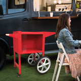 Carrello per popcorn KuKoo rosso brillante con ruote grigie, posizionato accanto a una donna seduta, in un ambiente all'aperto vivace