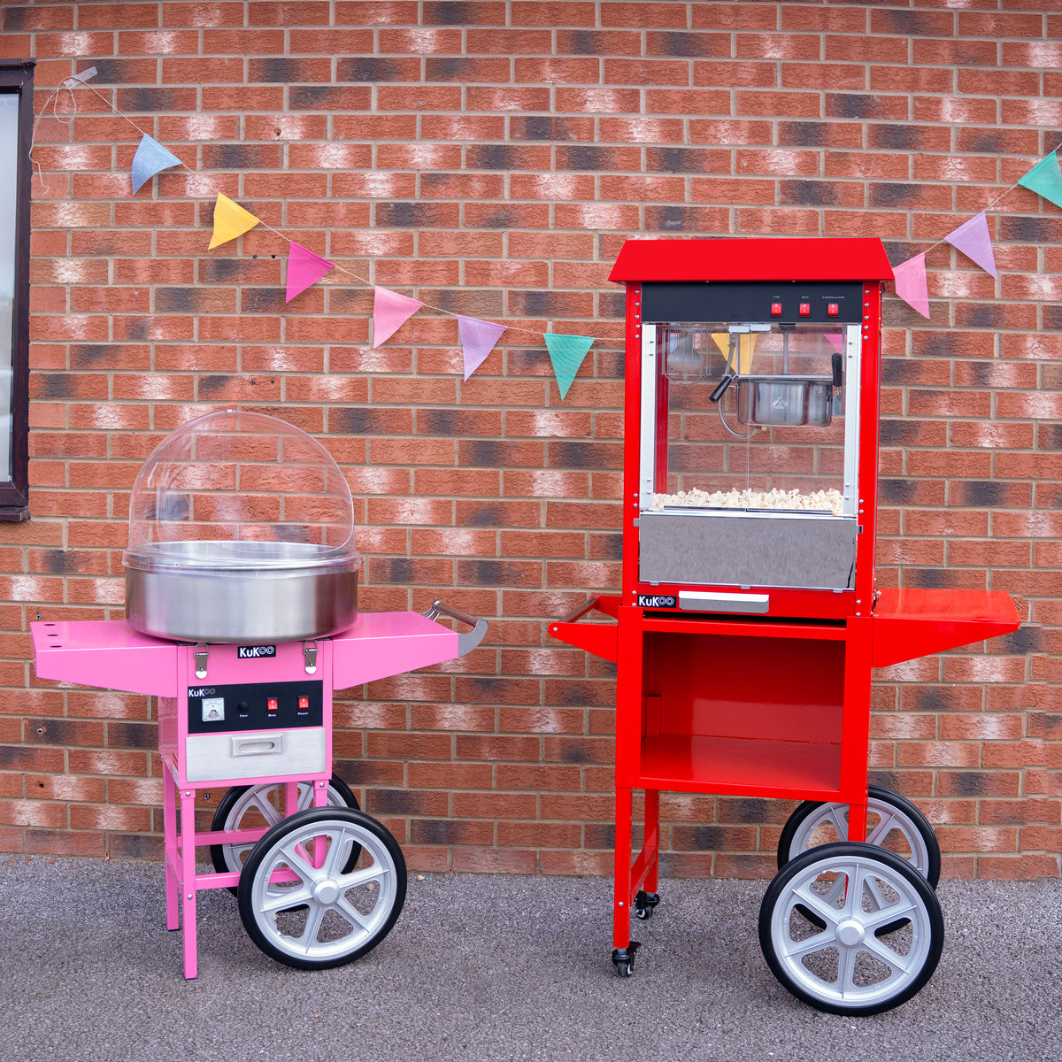 Carrello per popcorn rosso accanto a una macchina per zucchero filato rosa, entrambi su ruote, con bandierine colorate sullo sfondo di mattoni