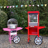 Carrello per popcorn rosso e macchina per zucchero filato rosa, entrambi con ruote, in un giardino verde con bandierine colorate