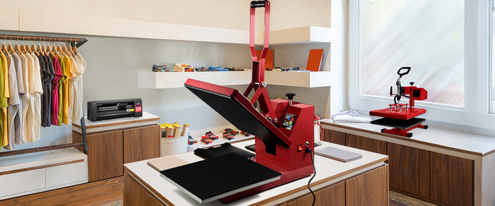 Pressa per trasferimento termico rossa su un tavolo di legno, con magliette colorate e attrezzature da stampa sullo sfondo
