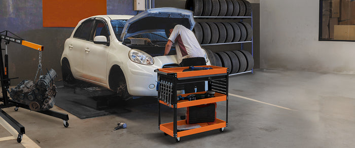 Carrello da officina arancione e nero con attrezzi, accanto a un'auto bianca con il cofano aperto in un ambiente di lavoro automobilistico